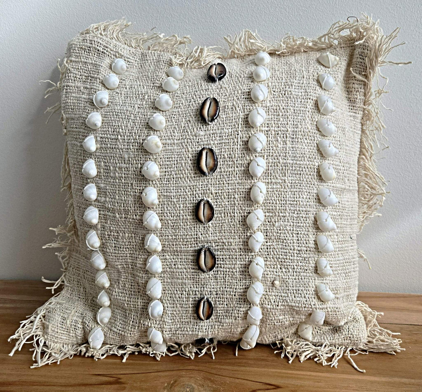 Coussin en tissu naturel orné de coquillages blancs et noirs, bordures frangées, style bohème.