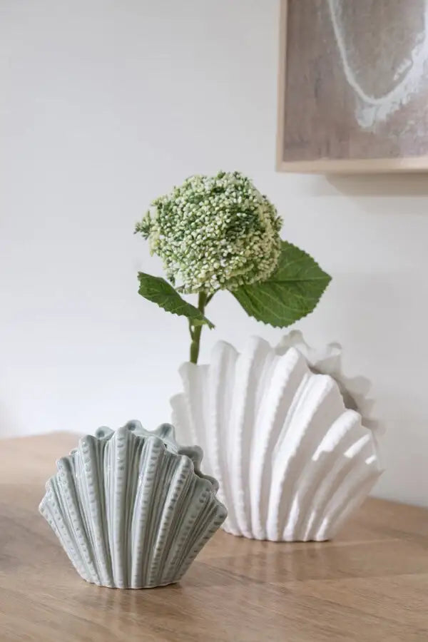 Deux vases en céramique forme coquillage, l'un blanc l'autre gris, avec fleur verte sur table en bois