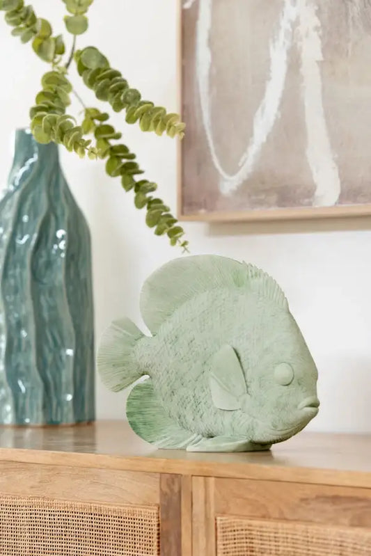 Statuette de poisson en céramique vert clair sur meuble en bois, avec vase bleu et plante en arrière-plan.